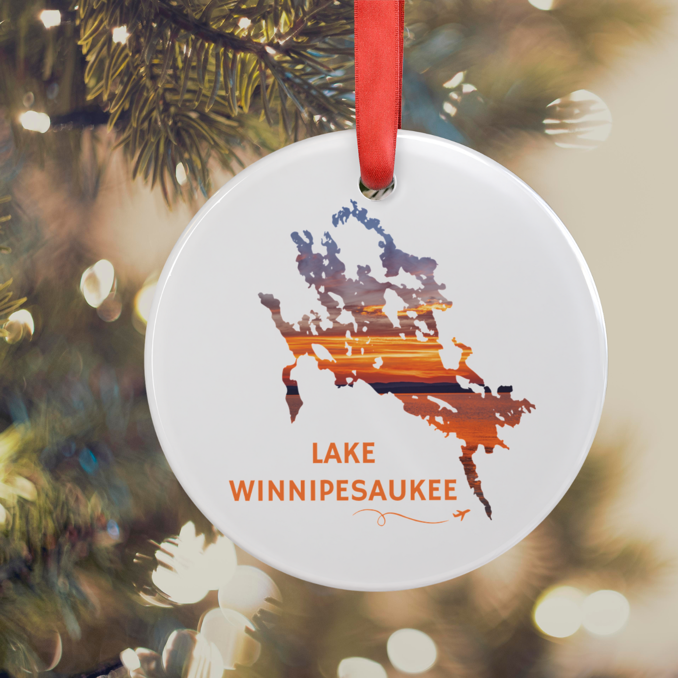 Round acrylic Lake Winnipesaukee ornament featuring a hand-drawn outline filled with a real sunset photograph, hanging from a Christmas tree with a red satin ribbon and glowing holiday lights in the background.