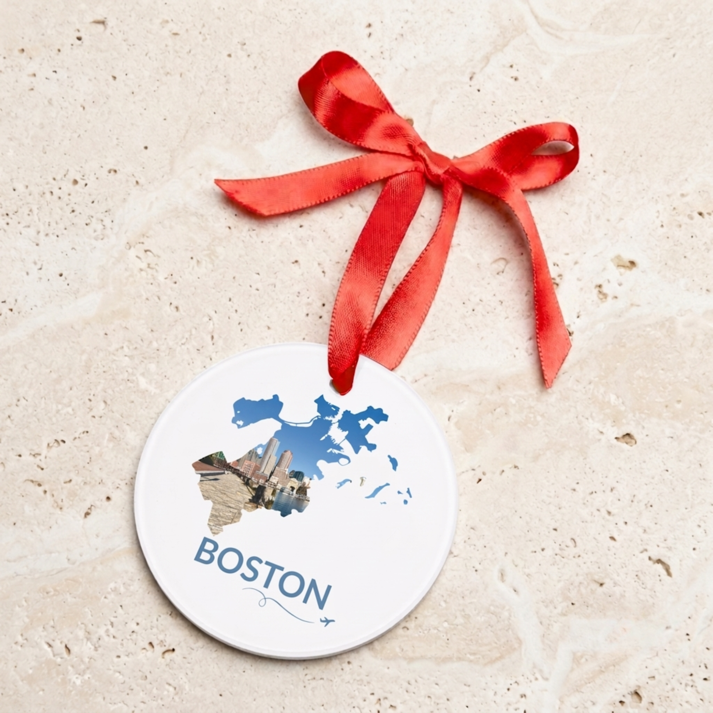 Round acrylic Boston ornament featuring a hand-drawn outline filled with a waterfront skyline photograph, hanging from a red satin ribbon against a neutral stone background.