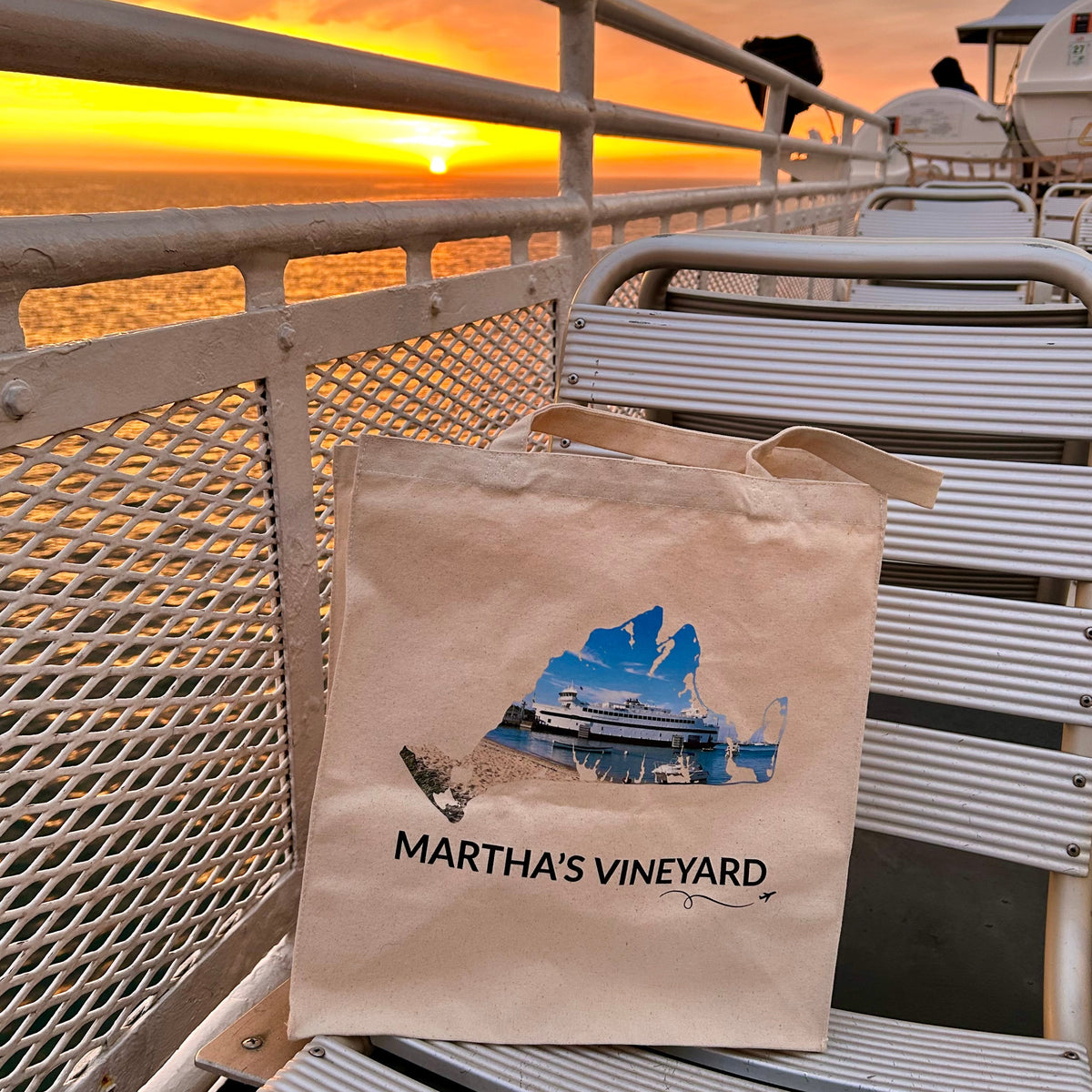 A beige canvas tote bag printed with a picture of the Steamship ferry docked in Vineyard Haven, MA, inside of the Martha's Vineyard outline, and the text 'MARTHA'S VINEYARD' with a plane and swirly line sits on the metal seat of a boat deck during a vibrant sunrise over the water.