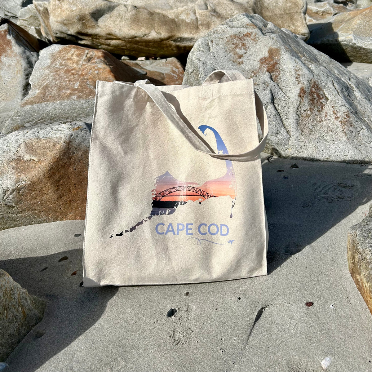 A beige canvas tote bag with a hand-traced Cape Cod map filled with an image of the Bourne Bridge at sunset, placed on a sandy beach among large rocks.