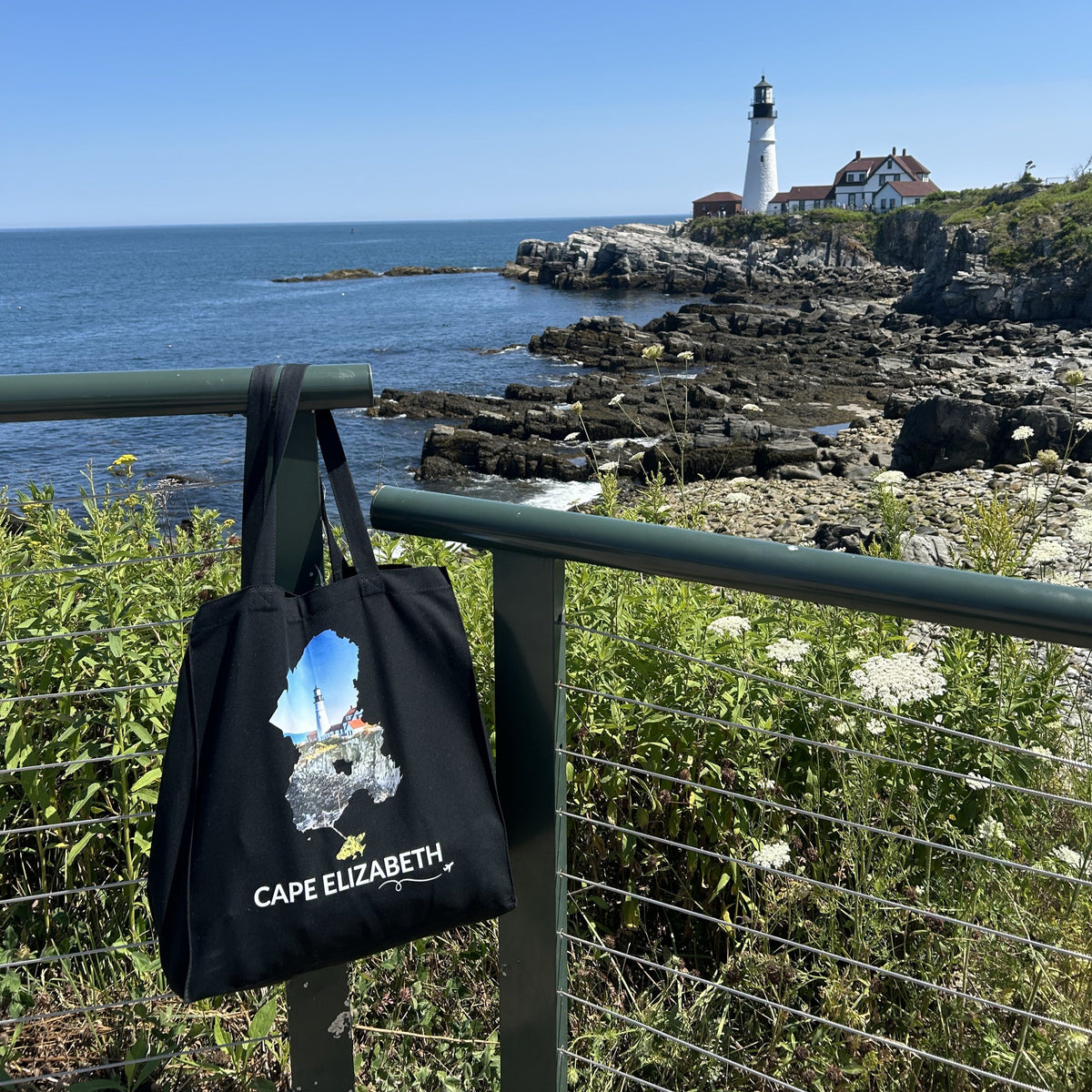 Black Cape Elizabeth tote bag displayed near Portland Head Light, featuring the lighthouse and rocky coastline inside the Cape Elizabeth outline, white Cape Elizabeth lettering, and a small airplane with swirl