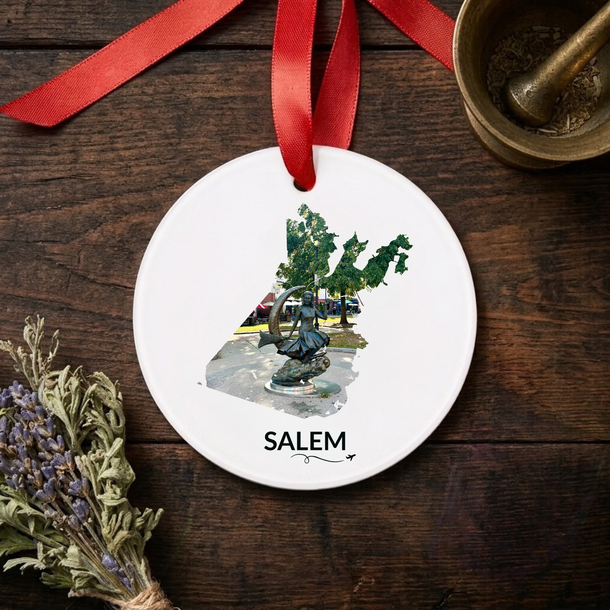 White Salem ornament with red satin bow, featuring the Bewitched Sculpture inside the Salem outline, SALEM lettering, and a small airplane with swirl, displayed on a dark wooden table beside dried lavender.