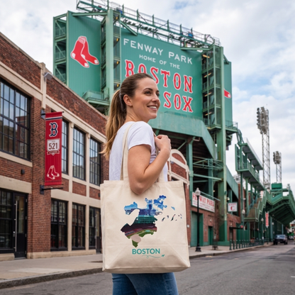 Woman carrying a natural cotton canvas tote bag featuring a hand-drawn Boston outline filled with an aerial view of Fenway Park while standing outside the iconic ballpark. The word "Boston" appears below the artwork with a small airplane icon and looping flight path.