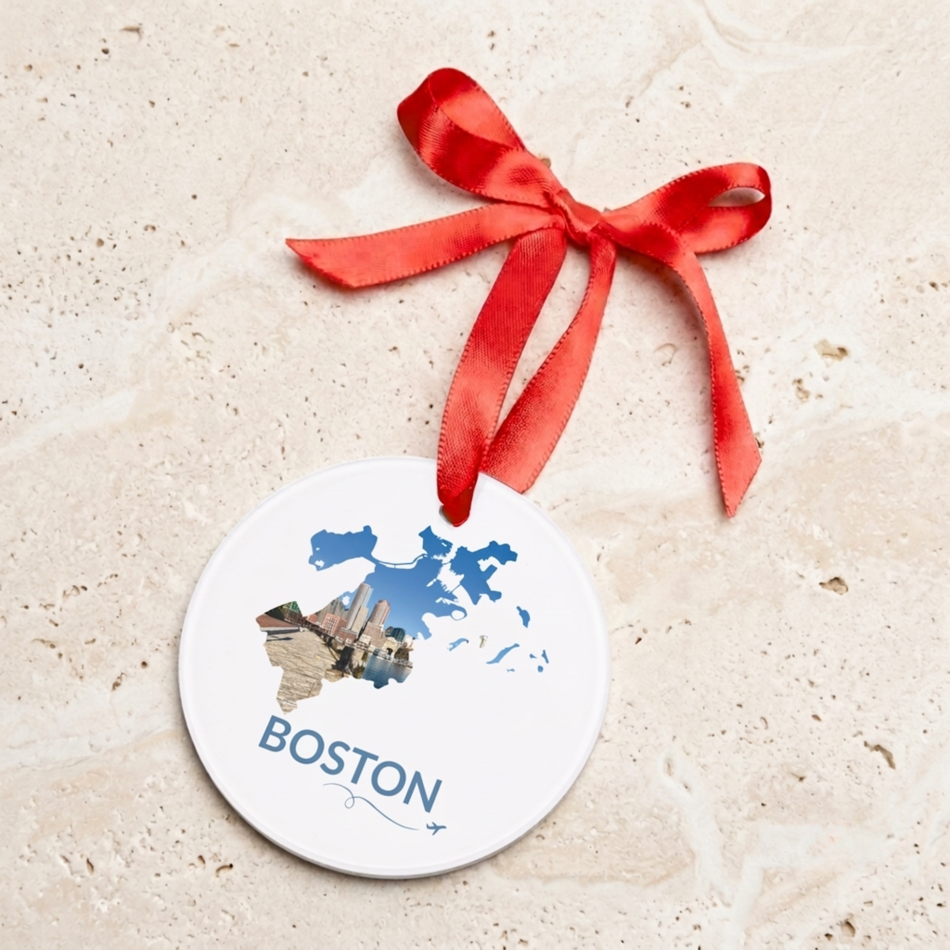 Round acrylic Boston ornament featuring a hand-drawn outline filled with a waterfront skyline photograph, hanging from a red satin ribbon against a neutral stone background.