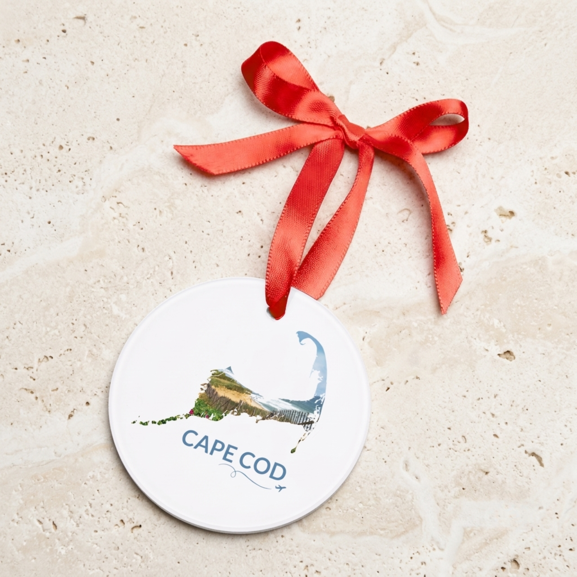 Round ceramic Cape Cod ornament featuring a hand-drawn outline filled with beach roses, sand fencing, and the Atlantic coastline, displayed with a red satin ribbon.