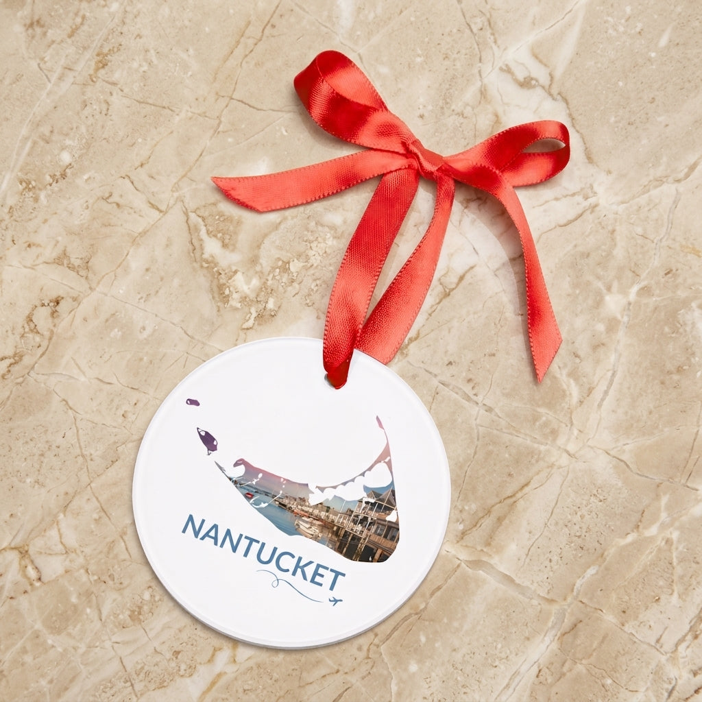 White Nantucket ornament with red ribbon resting on a stone surface, featuring a sunset harbor scene inside the island outline, blue Nantucket lettering, and airplane swirl