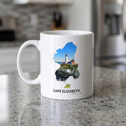 White Cape Elizabeth mug on a kitchen countertop featuring Portland Head Light and the rocky Maine coastline inside the Cape Elizabeth outline, black Cape Elizabeth lettering, and a small airplane with swirl