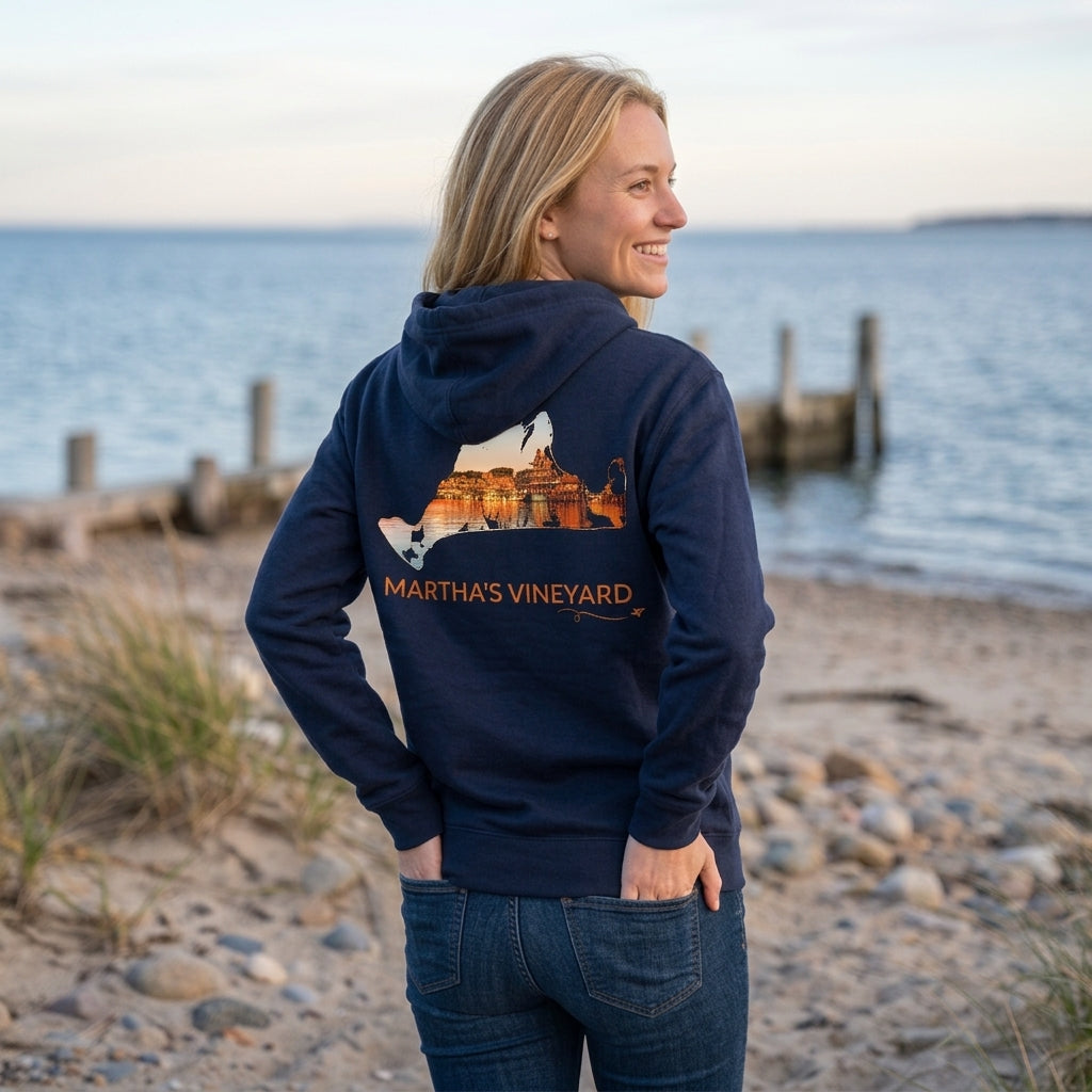 Woman wearing a navy Martha's Vineyard zip-up hoodie by the water, showing Oak Bluffs Harbor at sunset inside a hand-traced island outline on the back