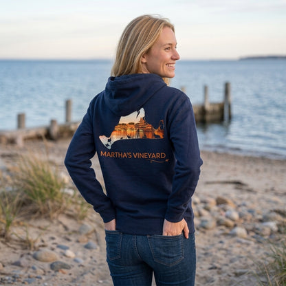 Woman wearing a navy Martha's Vineyard zip-up hoodie by the water, showing Oak Bluffs Harbor at sunset inside a hand-traced island outline on the back