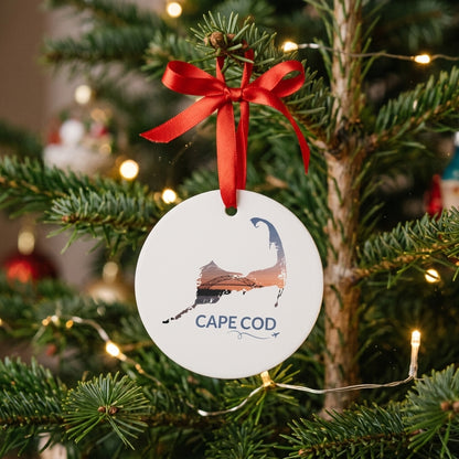 White Cape Cod ornament hanging on a Christmas tree with a red ribbon, featuring the Bourne Bridge at sunset inside the Cape Cod outline, Cape Cod lettering, and a small airplane with swirl