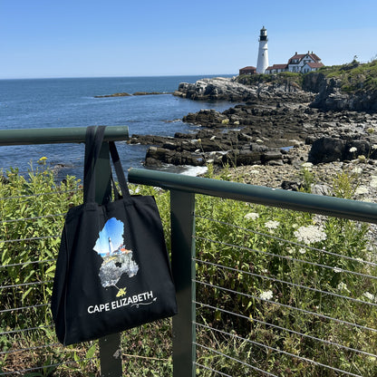 Black Cape Elizabeth tote bag displayed near Portland Head Light, featuring the lighthouse and rocky coastline inside the Cape Elizabeth outline, white Cape Elizabeth lettering, and a small airplane with swirl