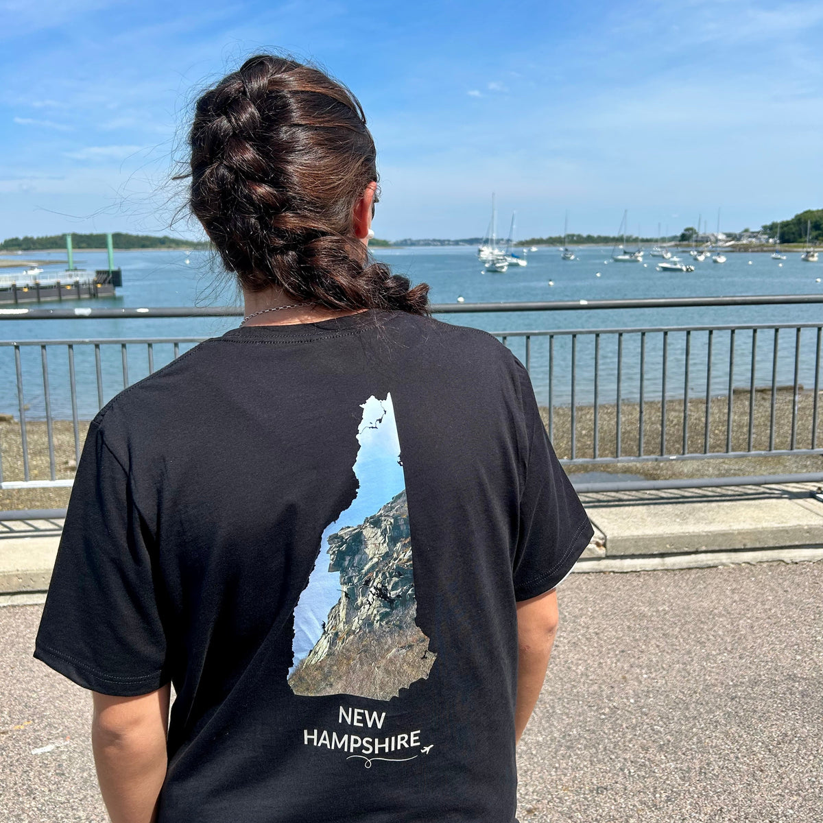 Person wearing a black New Hampshire T-shirt featuring the Old Man of the Mountain inside the state outline, with New Hampshire lettering and a small airplane with swirl, standing by a harbor with sailboats