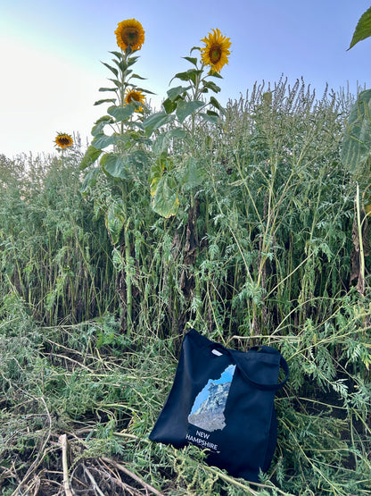 Black New Hampshire tote bag displayed among tall sunflowers, featuring the Old Man of the Mountain inside the New Hampshire outline, white New Hampshire lettering, and a small airplane with swir