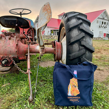 Navy Kancamagus Highway tote bag displayed beside a vintage tractor, featuring colorful fall foliage inside the New Hampshire outline, orange Kancamagus Highway lettering, and a small airplane with swirl