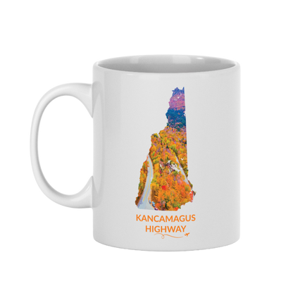White 11 oz ceramic mug featuring a hand-drawn New Hampshire outline filled with a photograph of the Kancamagus Highway winding through vibrant fall foliage. The words "Kancamagus Highway" appear below the artwork with a small airplane icon and looping flight path.