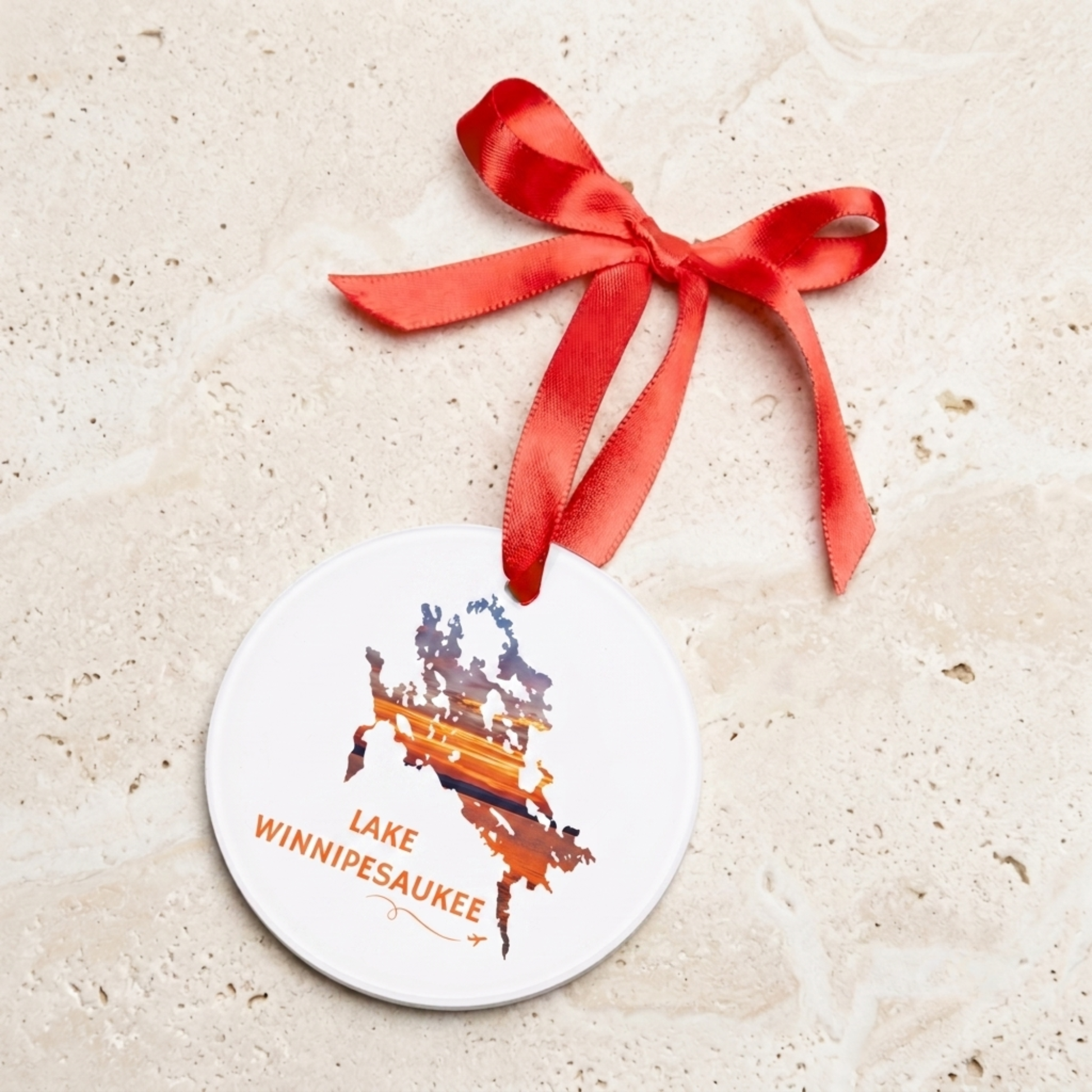 Lake Winnipesaukee acrylic ornament featuring a sunset lake scene displayed with a red satin ribbon against a neutral stone background.
