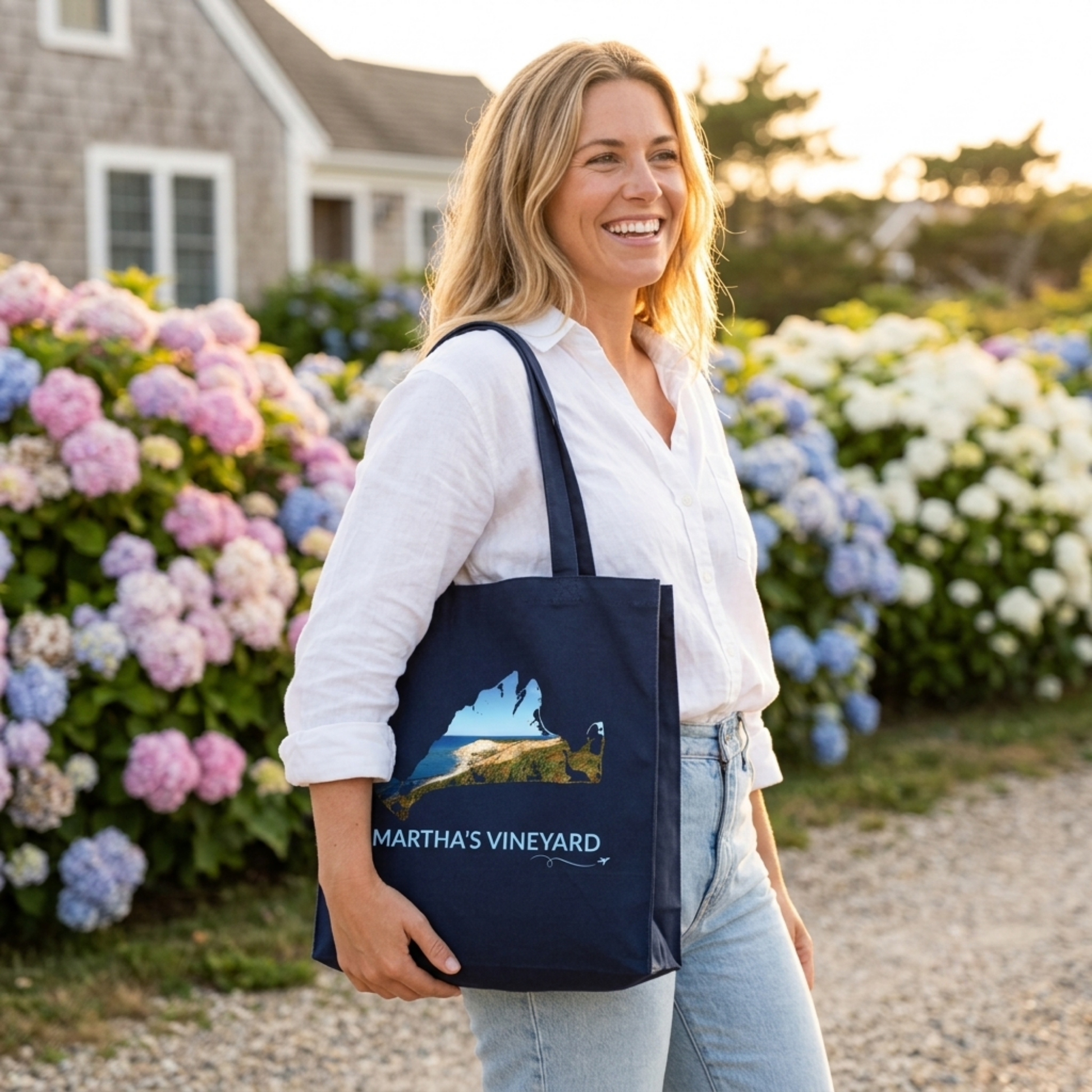 Woman carrying a navy Martha's Vineyard cotton canvas tote bag featuring a hand-drawn island outline filled with the Aquinnah Cliffs and Atlantic Ocean. The words "Martha's Vineyard" appear below the artwork with a small airplane icon and looping flight path.