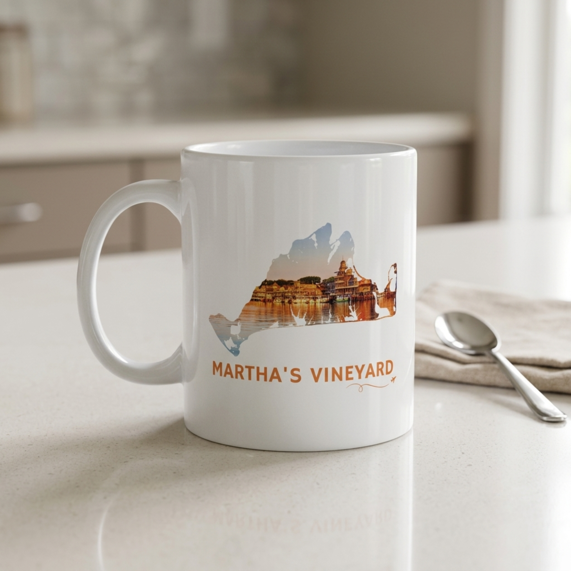 White ceramic Martha's Vineyard mug featuring a hand-drawn island outline filled with a sunset view of Oak Bluffs Harbor. "Martha's Vineyard" appears below the artwork with a small airplane icon and looping flight path. Displayed on a kitchen counter beside a spoon.