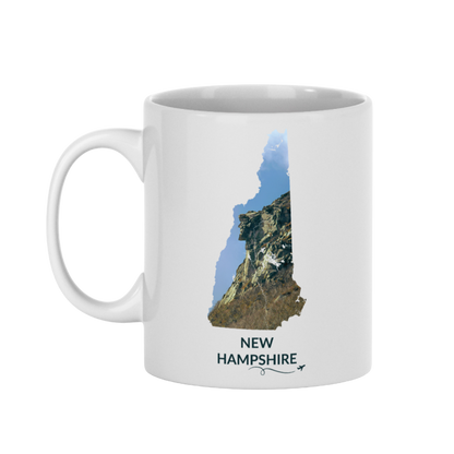 White 11 oz ceramic mug featuring a hand-drawn New Hampshire outline filled with a photograph of the Old Man of the Mountain. The words “New Hampshire” appear below the artwork with a small airplane icon and looping flight path.