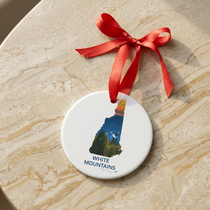 Round New Hampshire ornament featuring a hand-drawn state outline filled with a sunset photograph of the White Mountains, displayed with a red satin ribbon on a neutral stone surface.