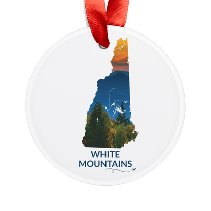 Round acrylic New Hampshire ornament featuring a hand-drawn state outline filled with a sunset photograph of the White Mountains. The words "White Mountains" appear below the artwork with a small airplane icon and looping flight path.