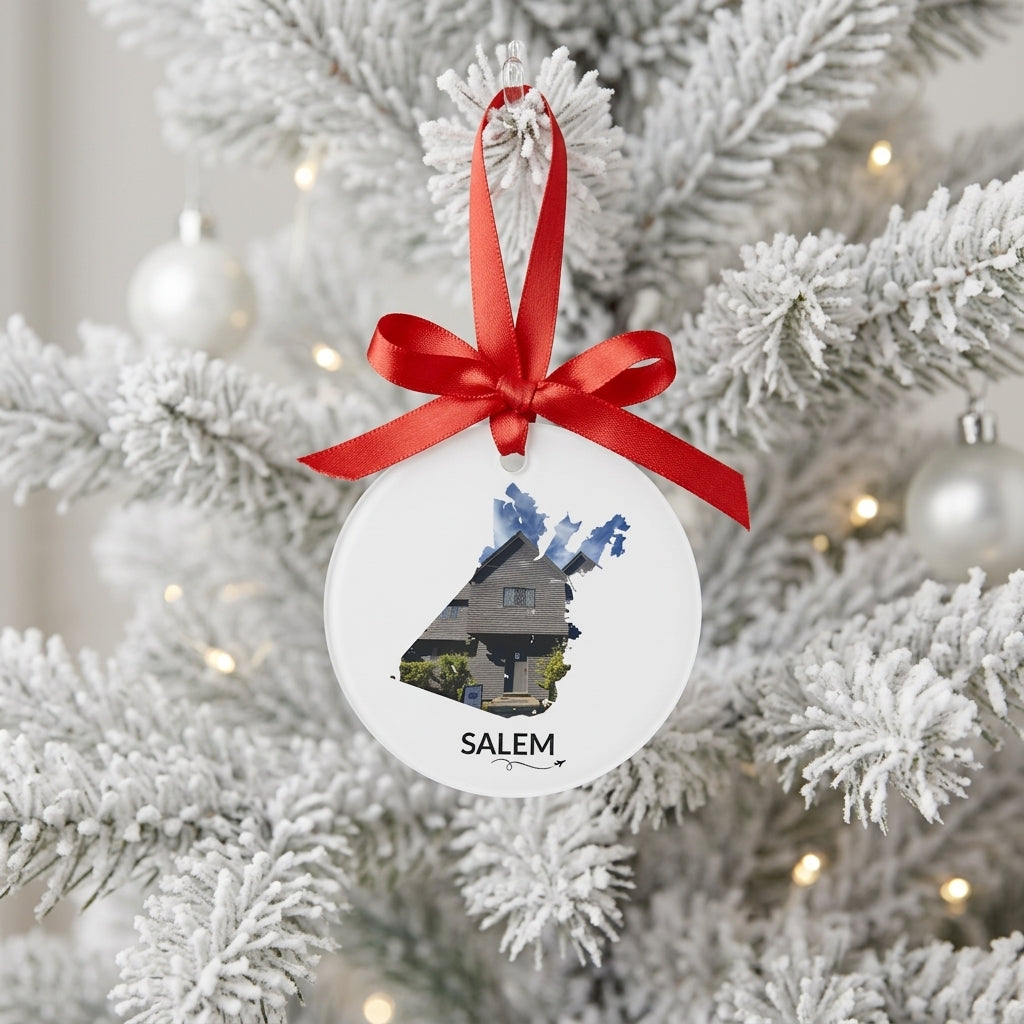 White Salem ornament hanging from a red satin bow on a snow-covered Christmas tree, featuring the Witch House inside the Salem outline, black SALEM lettering, and a small airplane with swirl.