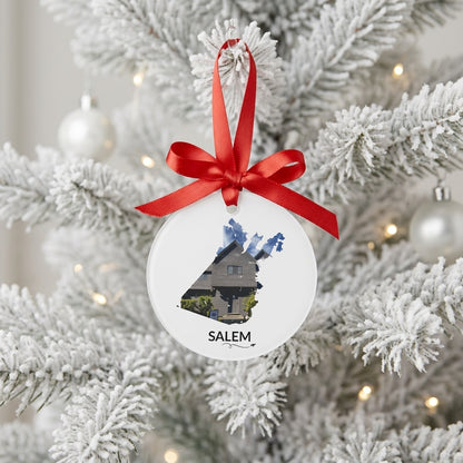 White Salem ornament hanging from a red satin bow on a snow-covered Christmas tree, featuring the Witch House inside the Salem outline, black SALEM lettering, and a small airplane with swirl.